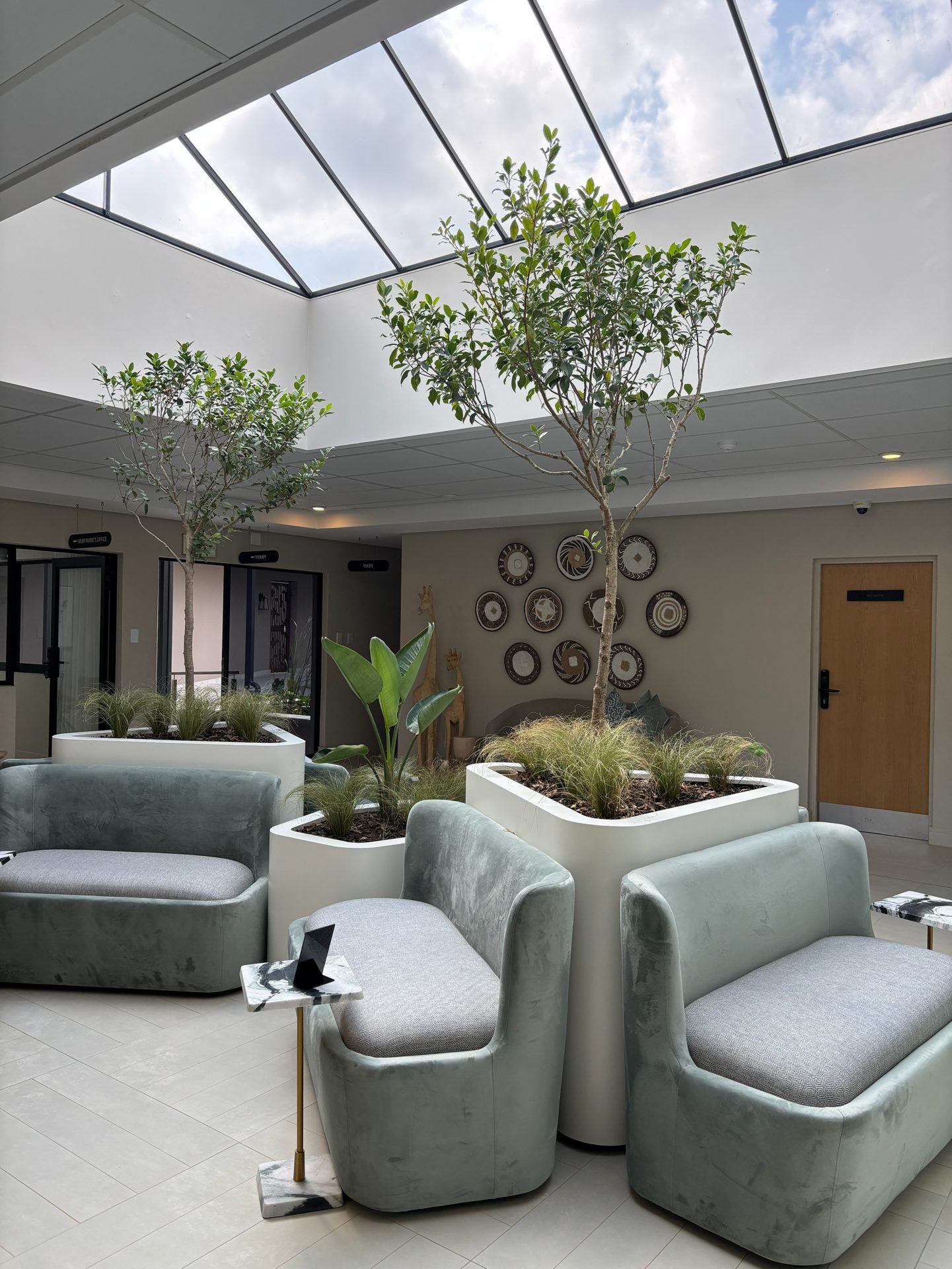 Atrium lounge at The Ark beneath a glazed skylight, with indoor trees and soft green seating